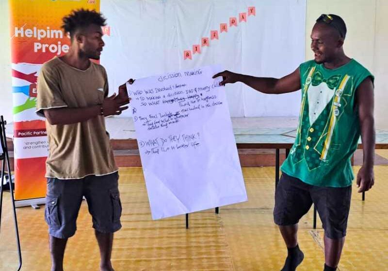 Two men hold up a large piece of paper with text on it. The title on the paper says 'decision making'. They are presenting in front of the Helpim Sista project pull up banner