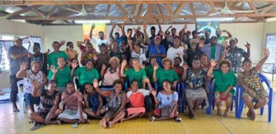 A large group of men and women sit inside together for a group photo. They are smiling and waving their hands.