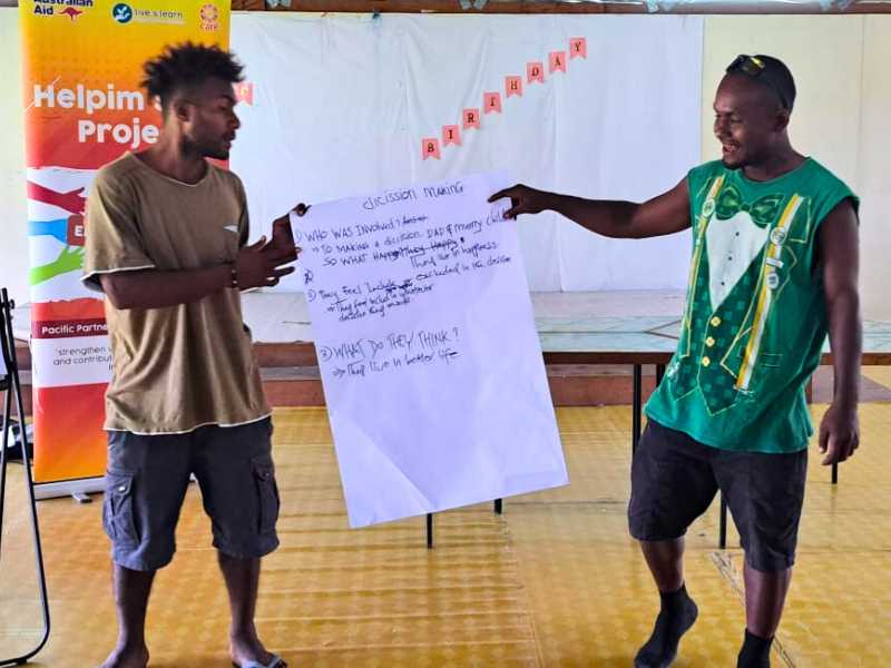 Two men hold up a large piece of paper with text on it. The title on the paper says 'decision making'. They are presenting in front of the Helpim Sista project pull up banner