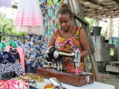 Lilian stands behind a new sewing machine, sewing clothes for her small business. 