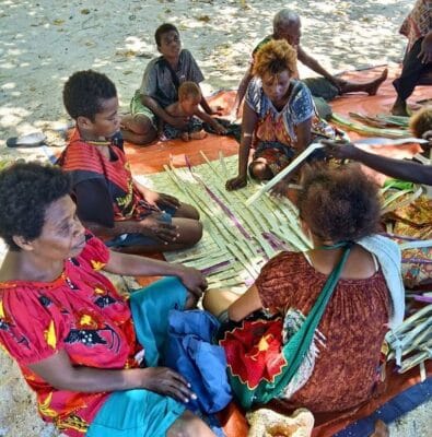 A group of women sit on the beach weaving a mat 